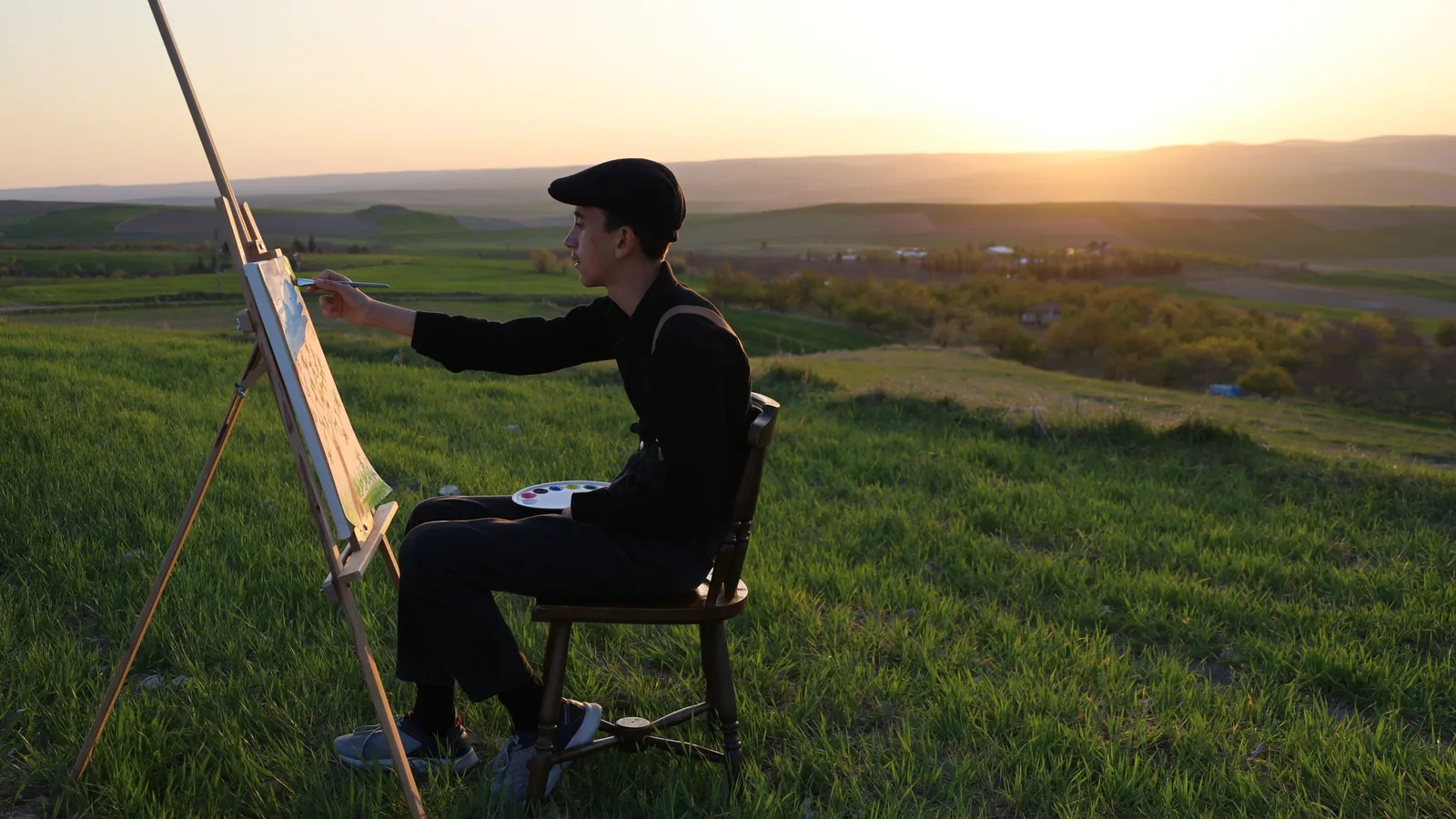 An artist painting at an easel on a hillside at sunset