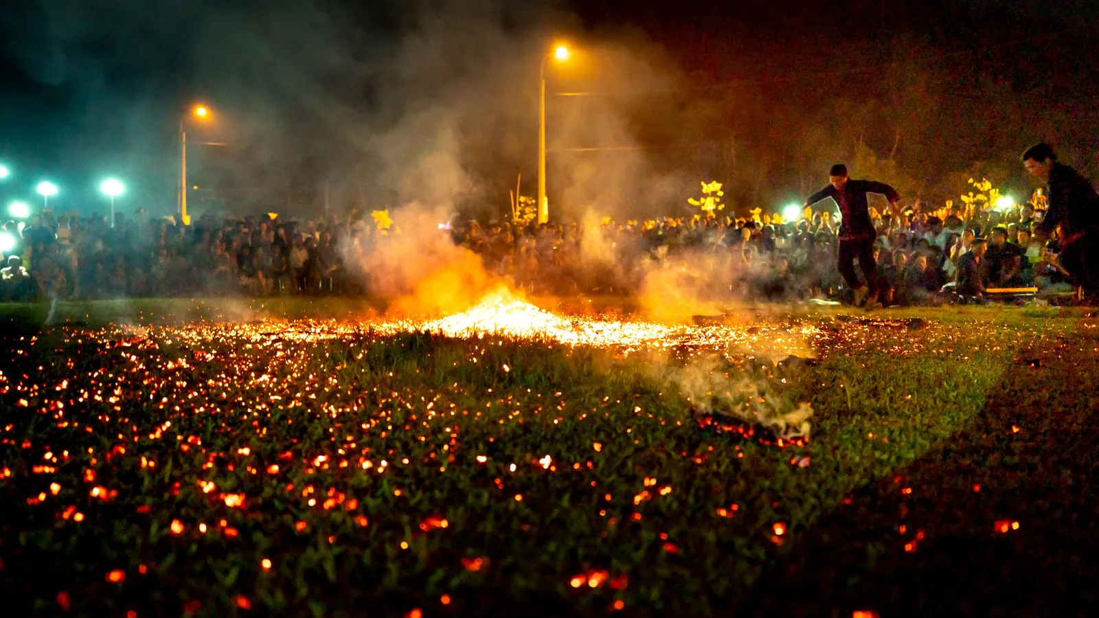 A field of glowing embers at night with people gathered around the fire