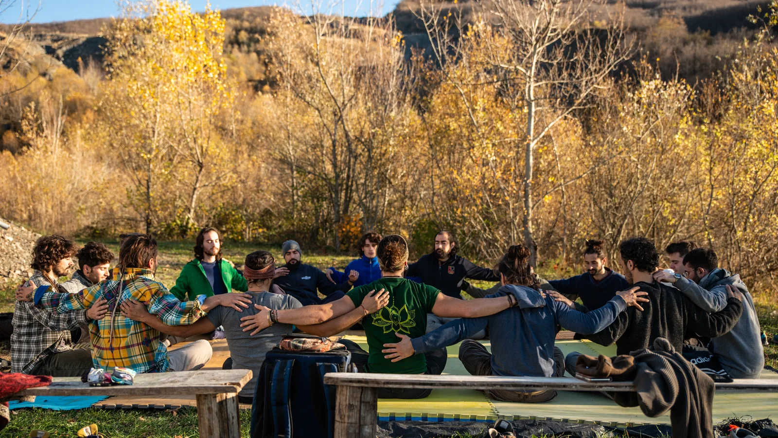 A circle of people sitting close together on benches under autumn trees, arms around each other