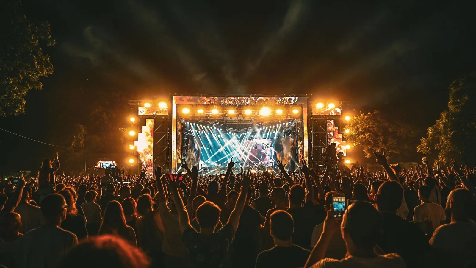 An outdoor festival stage under trees lit warm at night with a crowd in front