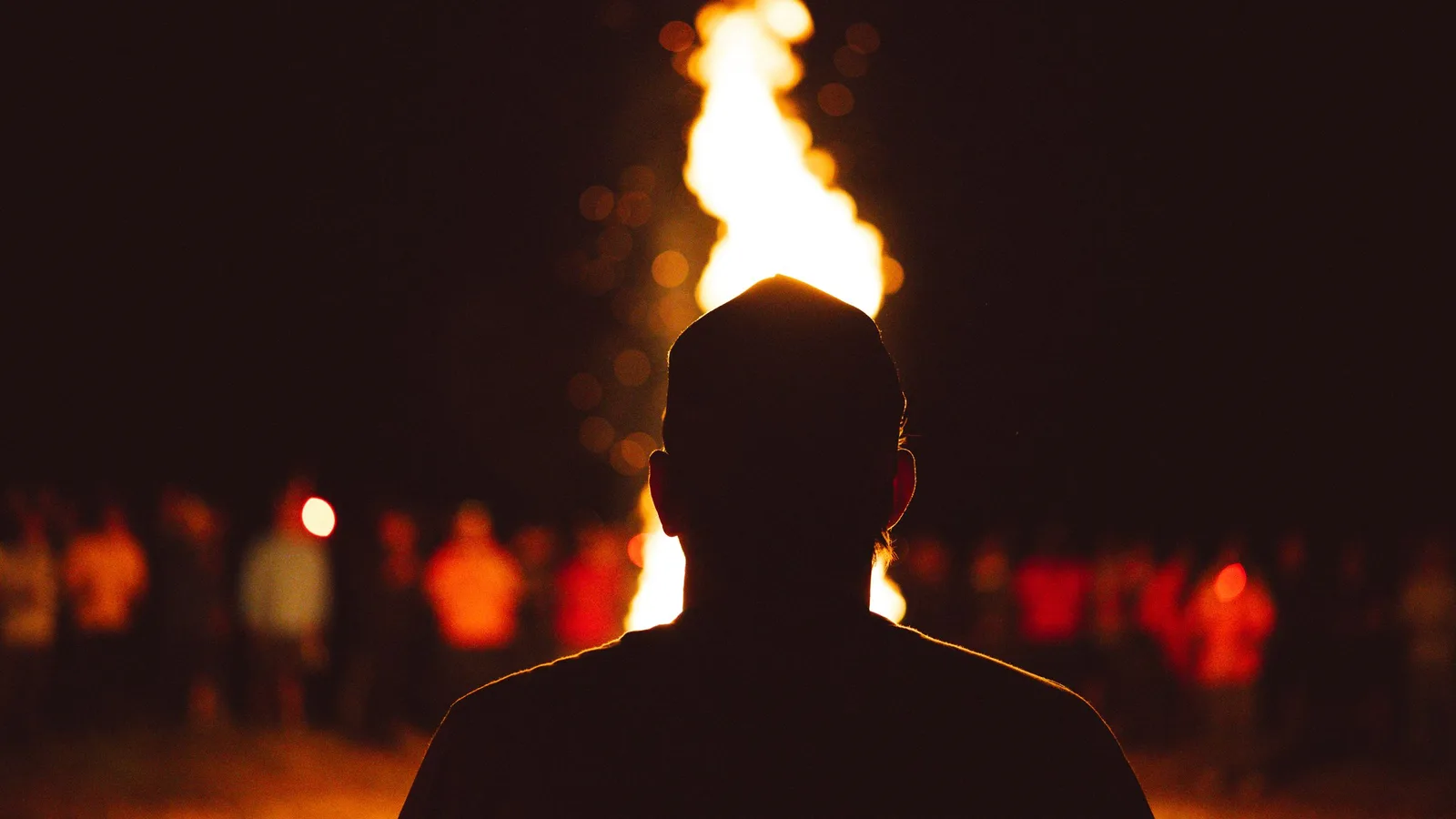 A silhouette facing a tall bonfire with a crowd gathered beyond