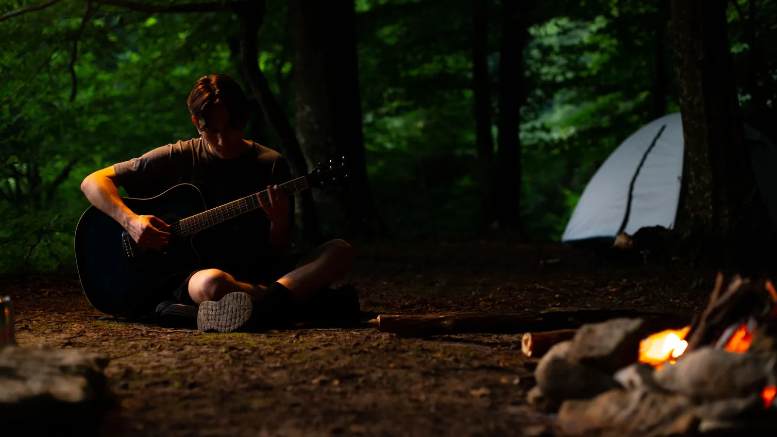 A person playing acoustic guitar by a campfire in the forest at night