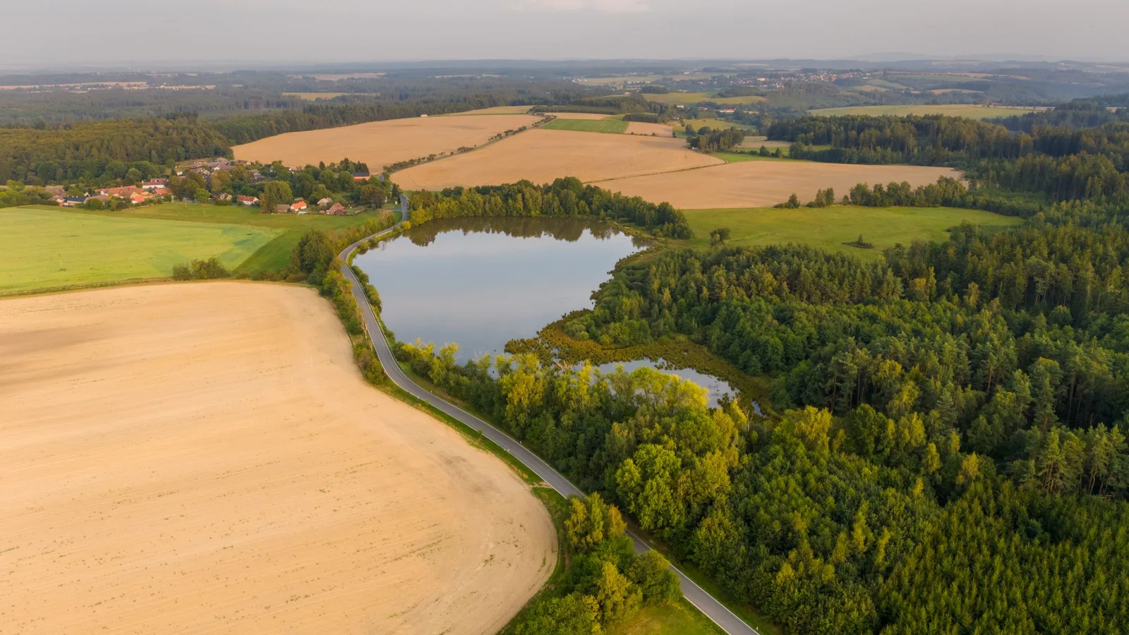 Aerial view of a small lake between fields, woods and a village